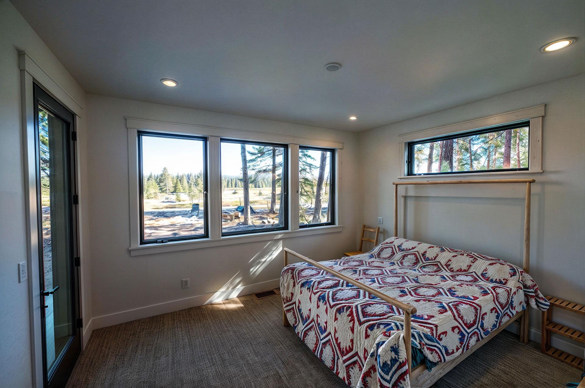 Bedroom with a queen bed, large picture windows, and a glass door overlooking a wooded landscape.