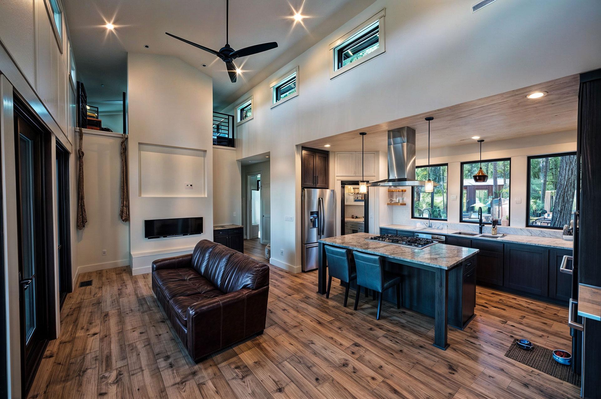 Open concept living area with a vaulted ceiling, fireplace, kitchen island, and large windows overlooking the outdoors.