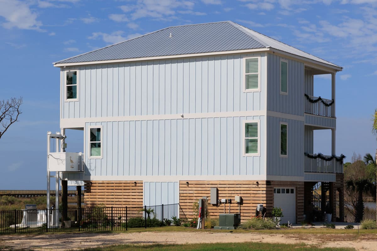 House plan exterior: two-story home with light blue vertical siding, metal roof, and multiple covered balconies. Elevated on pilings.