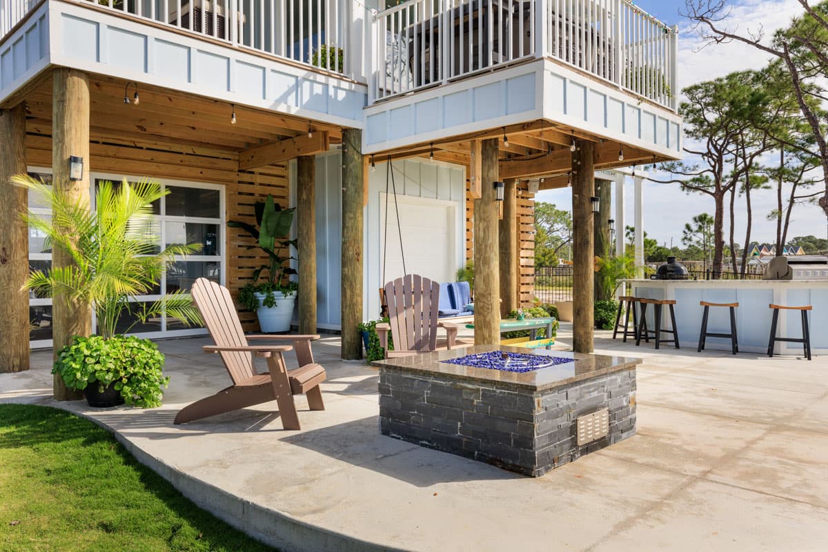House plan exterior featuring a raised structure on pilings, a deck, outdoor bar, fire pit, and Adirondack chairs.