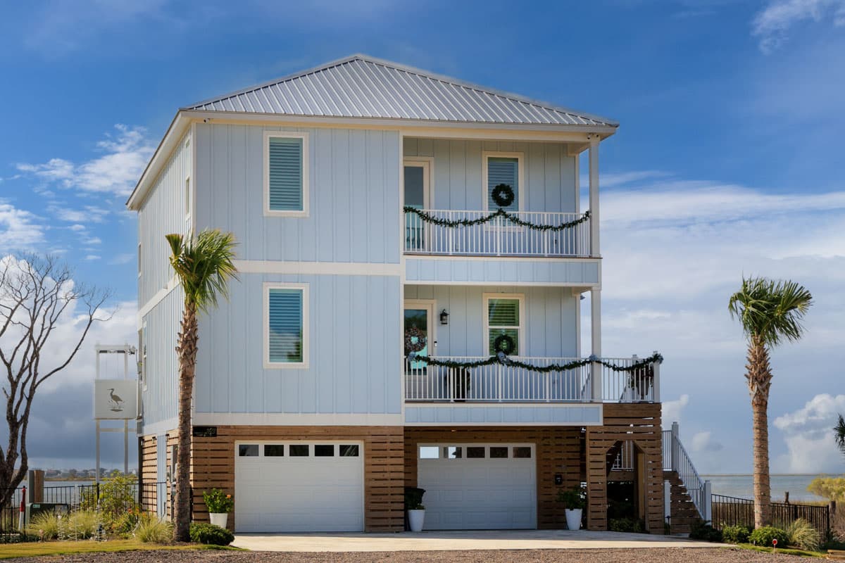 House plan exterior. Two-story coastal-style house with metal roof, two balconies with railing, and two-car garage at ground level.
