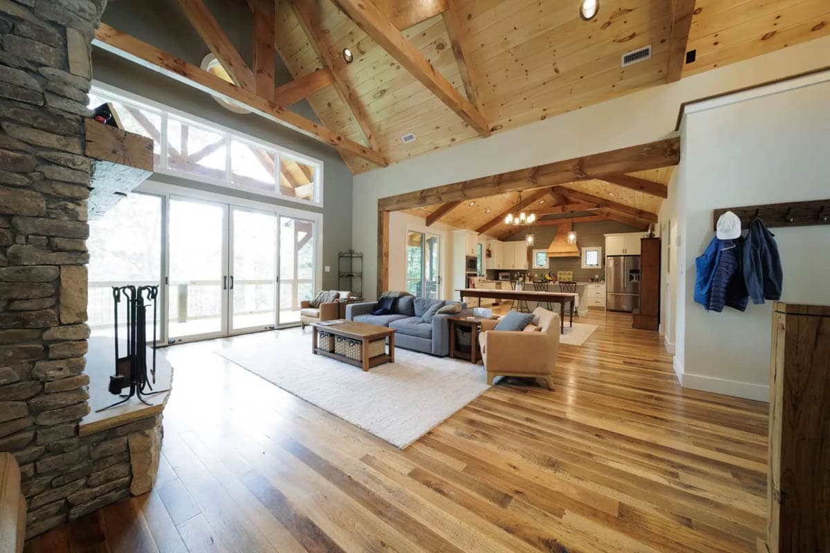 Great room interior with vaulted wood beam ceiling, stone fireplace, large windows, and open kitchen.