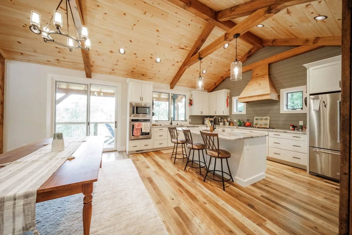 Interior view of open-concept kitchen with vaulted wood-paneled ceiling, exposed beams, island with seating, and white cabinetry.