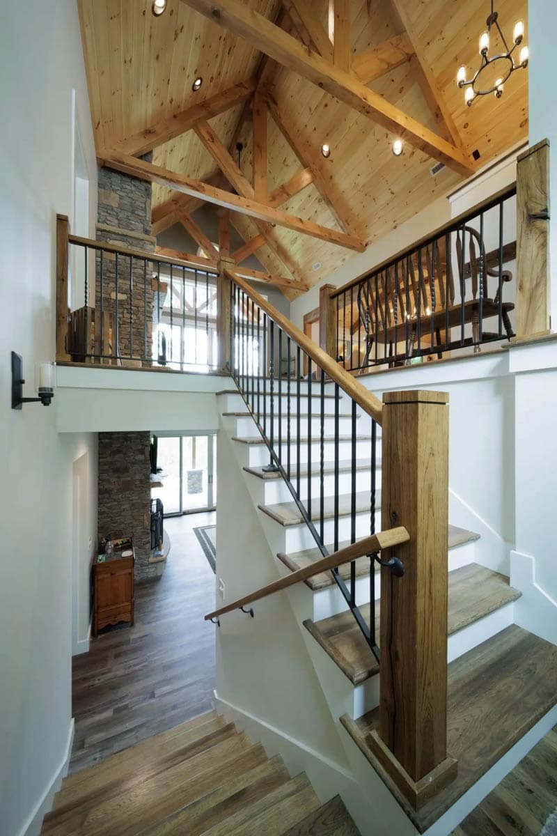 Interior view of a house with a grand staircase, vaulted wood-beamed ceiling, stone fireplace, and loft overlooking the living area.