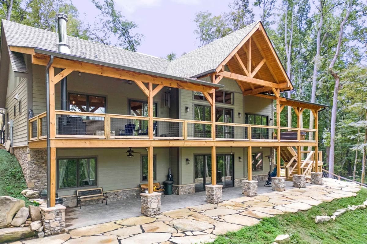 House plan exterior: Two-story home with a wraparound deck, stone foundation, and exposed timber framing.