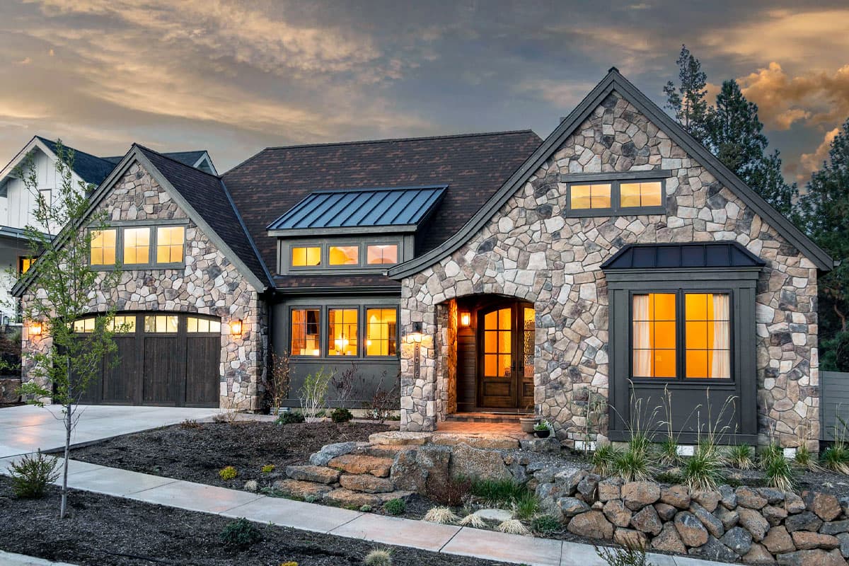 House plan exterior. Stone-clad, one-story home with gabled roof, dormers, and attached garage. Entry has a covered porch with stone steps.