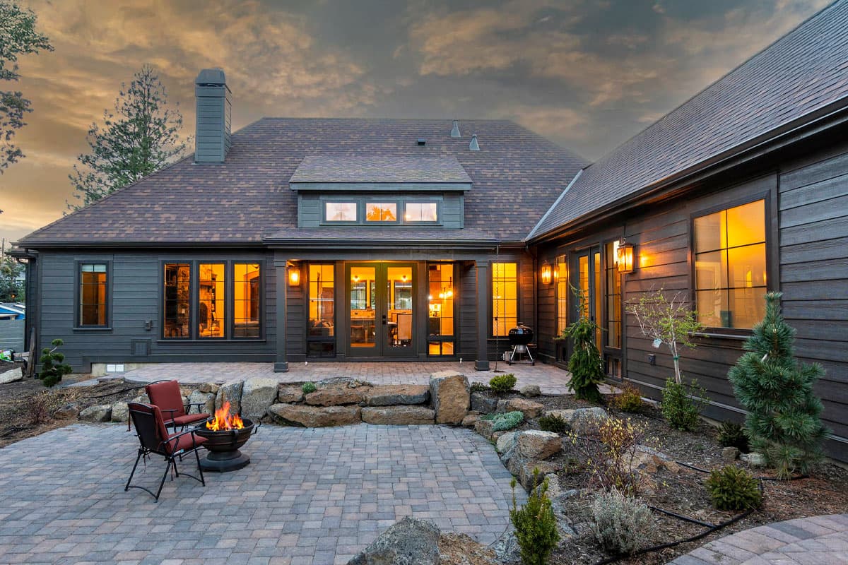 House plan exterior with dark siding, multiple gables, a prominent chimney, and a large stone patio with a fire pit.