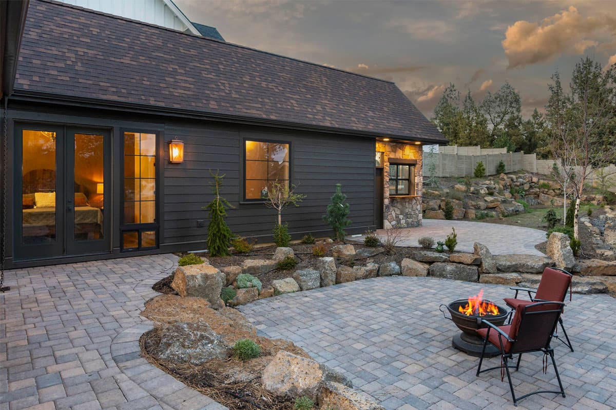House plan exterior with dark horizontal siding, stone accents, french doors, and a paved patio with fire pit.