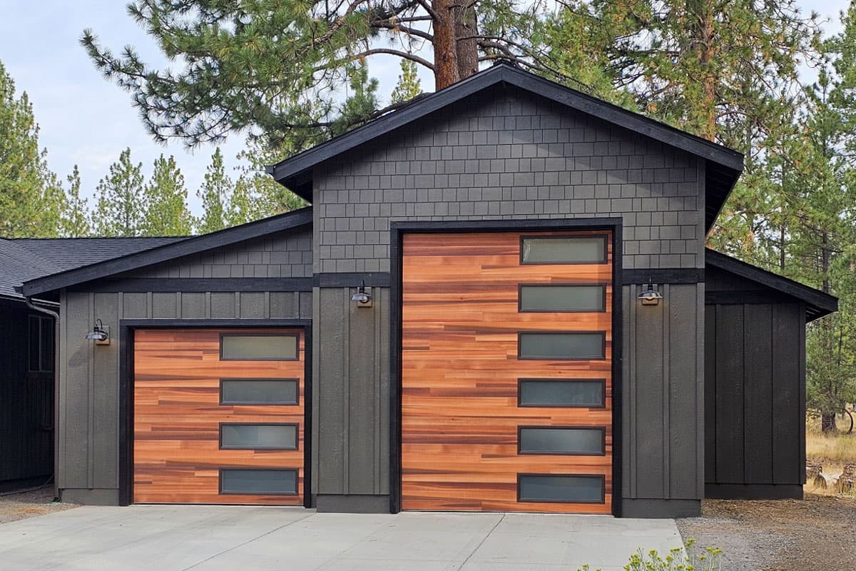 House plan exterior with two garage bays, wood-grain doors with rectangular windows, dark grey siding, and contrasting black trim.