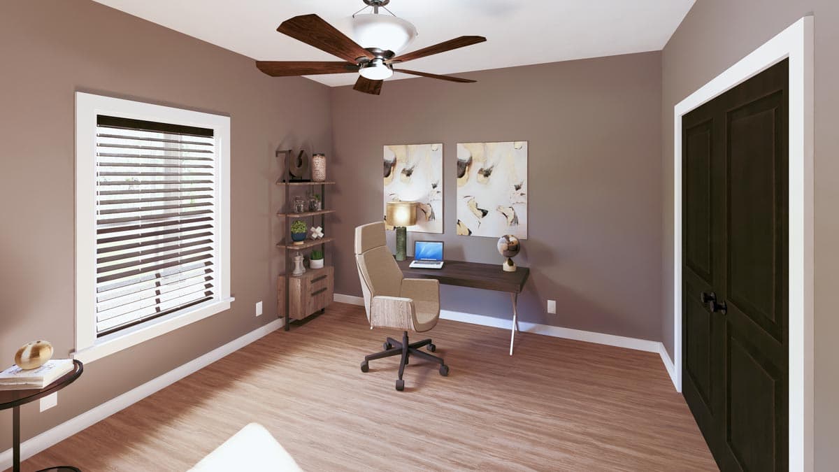 A modern home office with a desk, office chair, artwork, and a ceiling fan. Natural light streams in from a window with blinds.