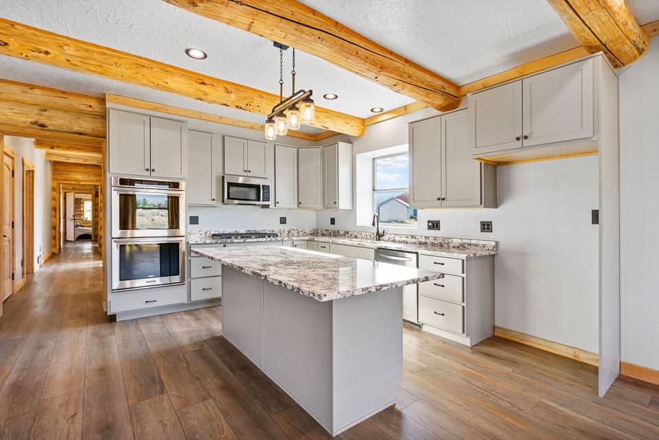 Kitchen interior featuring a central island, light gray cabinetry, stainless steel appliances, granite countertops, and exposed wood ceiling beams.