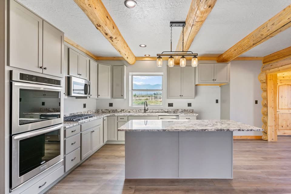 Kitchen with gray cabinets, granite counters, double ovens, island, and exposed wood ceiling beams.