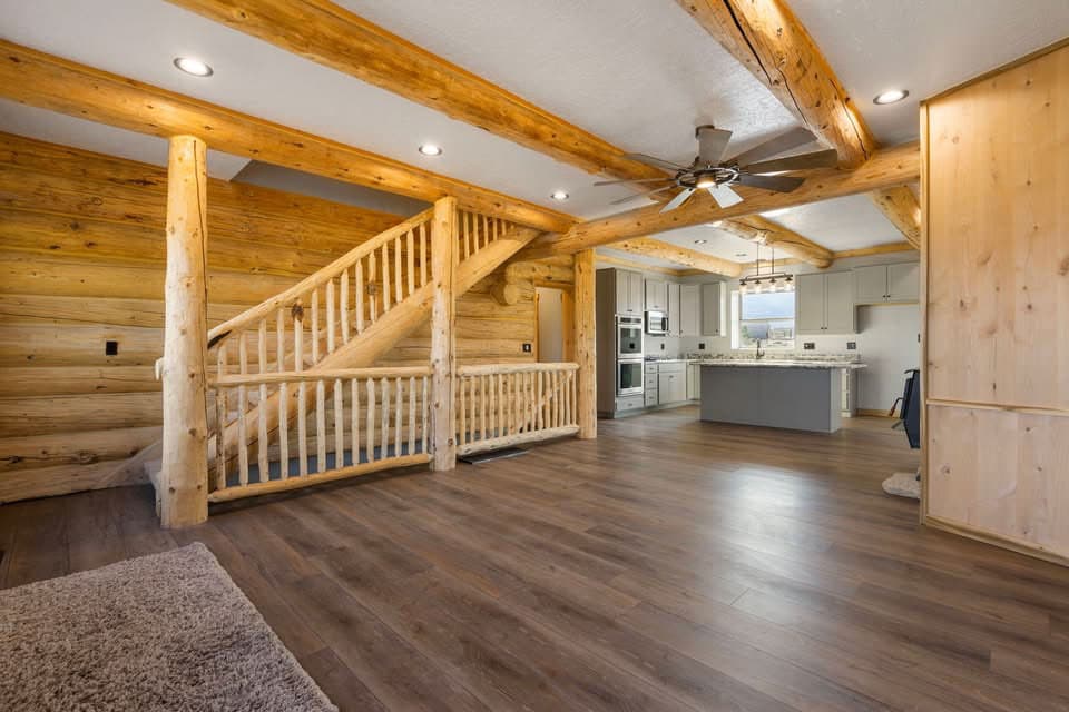 Log home interior with exposed wood beams, rustic staircase, and open-concept kitchen with island.