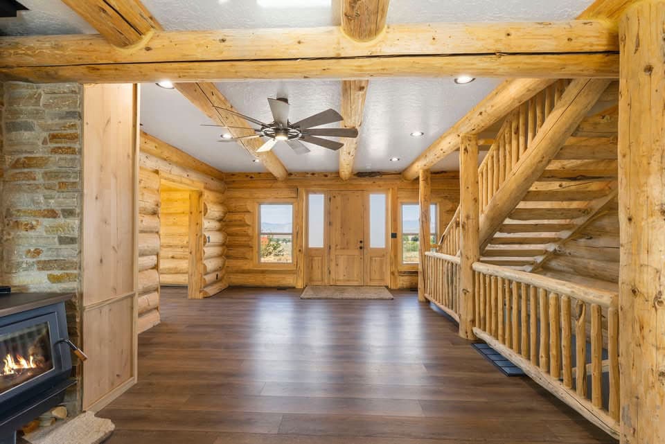 Log cabin interior with a stone fireplace, wood-paneled walls, and a staircase with a log railing leading upstairs.