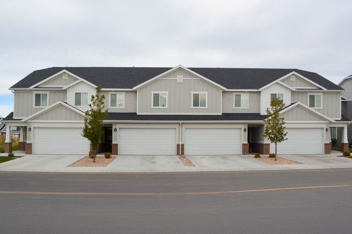 House plan exterior. Two-story townhouse with gabled roof, individual garages, and contrasting trim. Features include vertical siding and dormers.