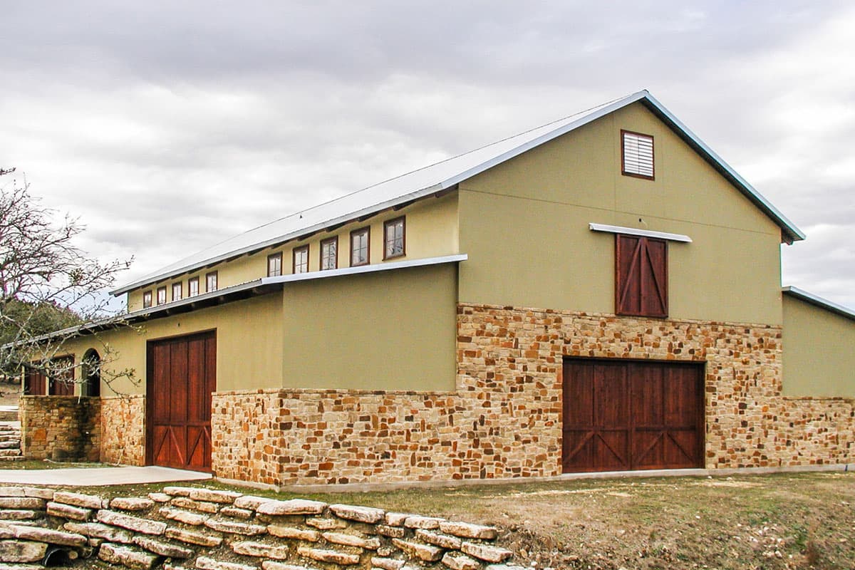 House plan exterior. Two-story structure with stone facade, metal roof, and large barn doors. Features include a gabled roof and small upper windows.