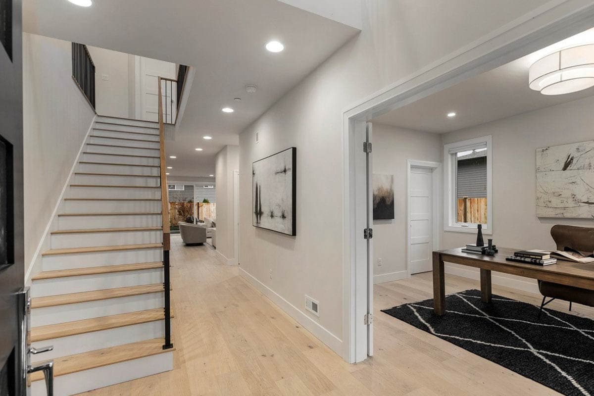 Interior view of a modern home's hallway with open-riser stairs and an adjacent office with a desk and minimalist art.