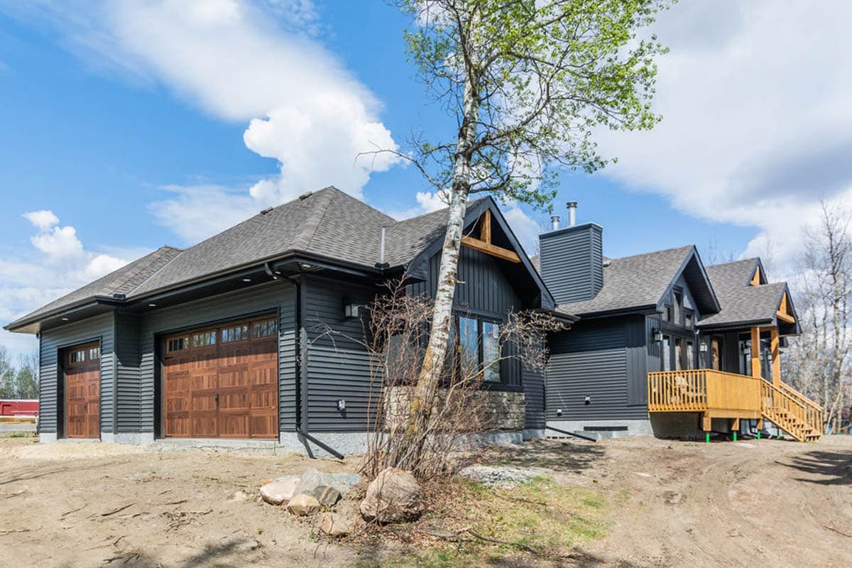 Modern house plan exterior with dark siding, wood garage doors, gables, stone accents, and a wood deck with stairs.