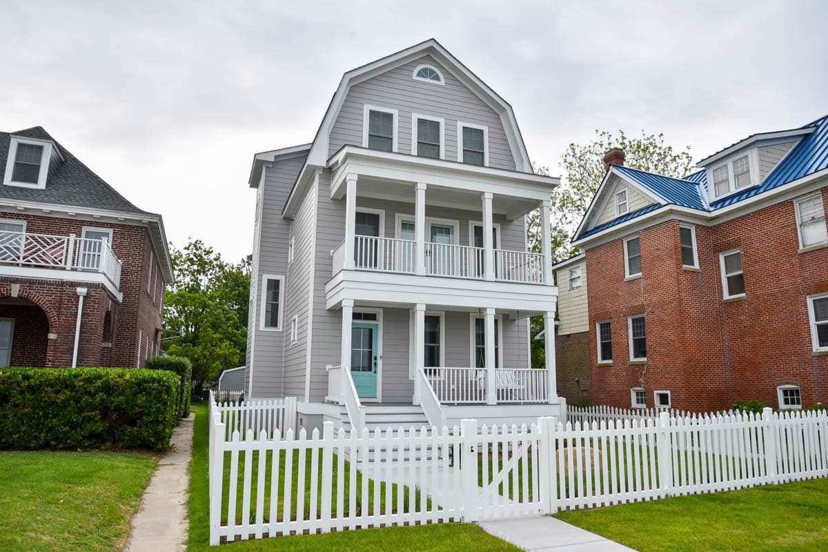 House plan exterior: Three-story gray house with double-level porches, gabled dormer, and white picket fence.