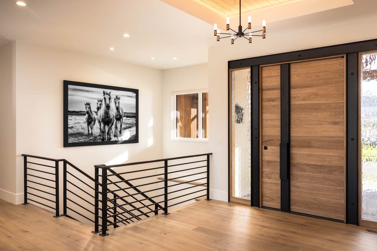 Interior view of a house entrance with wood doors, frosted glass sidelights, a black metal railing, and artwork of horses.