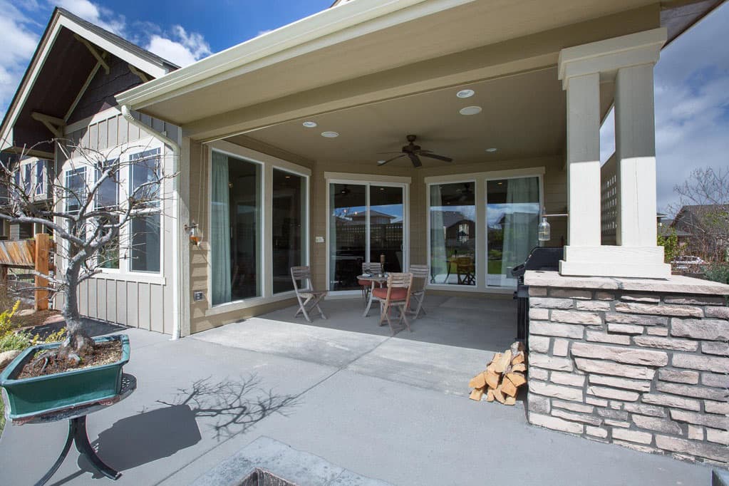 Covered patio with stone column, ceiling fan, outdoor dining set, and grill. House exterior features vertical siding and large windows.