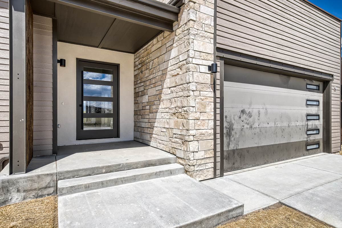 Modern house exterior showing a stone-clad corner, horizontal siding, a glass-paneled entry door, and a multi-panel garage door with windows.
