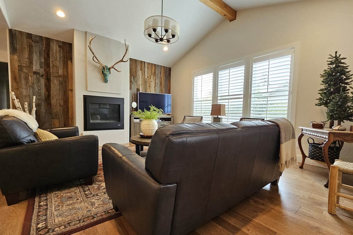 Living room with vaulted ceiling, wood accent walls, fireplace, leather sofas, and large windows with shutters.