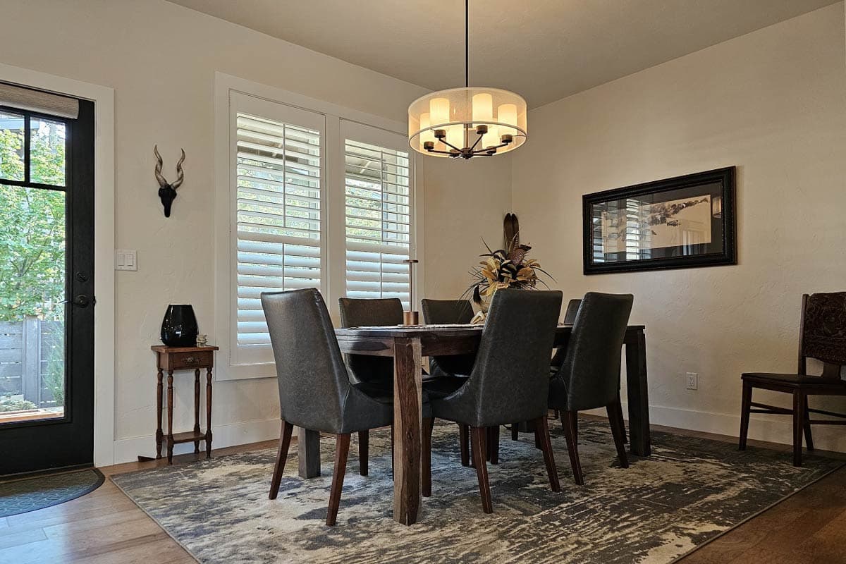 Dining room with wood table, upholstered chairs, pendant light, and large windows with shutters.