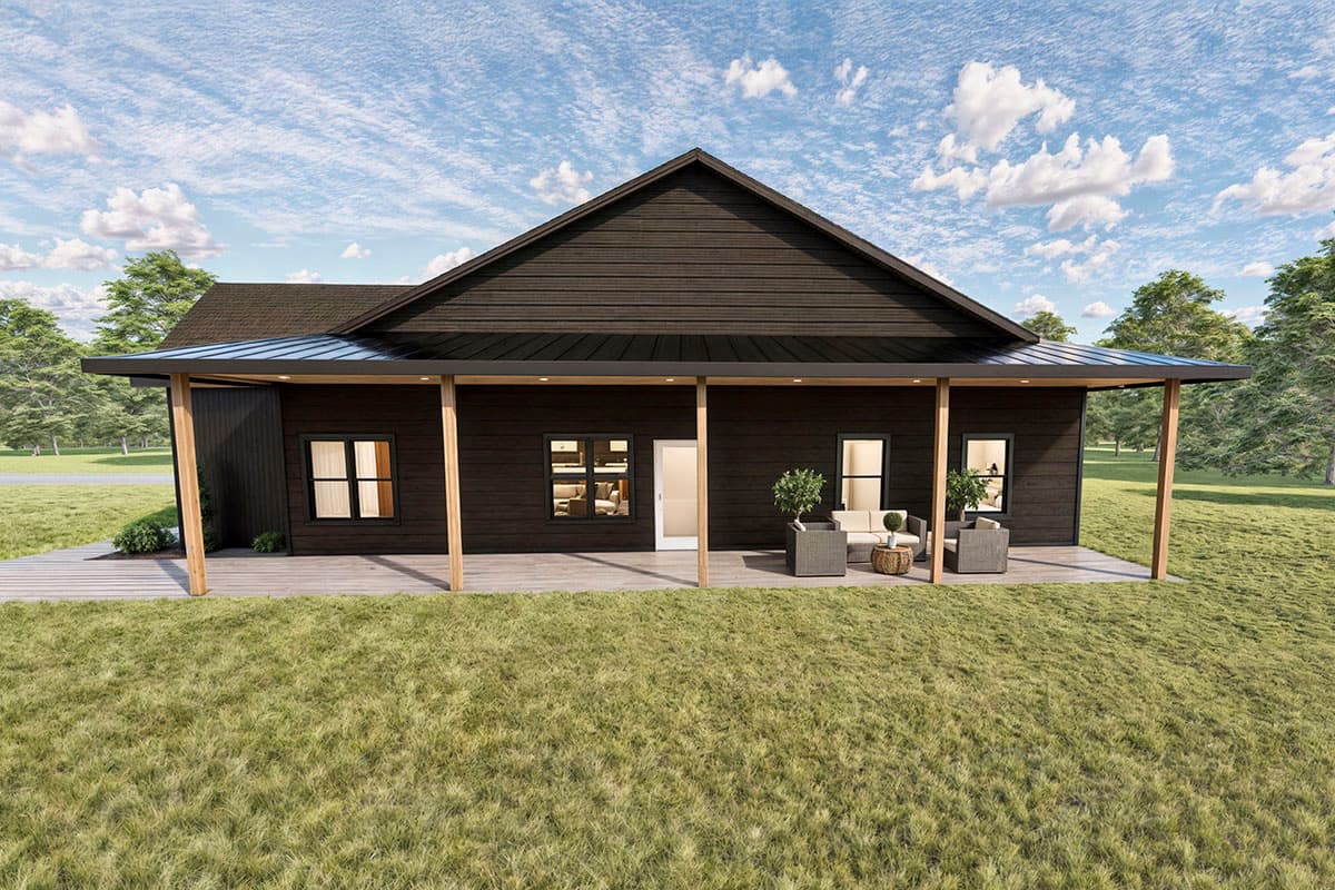A modern, dark-wood home with a covered porch and outdoor seating faces a grassy lawn under a blue sky dotted with clouds.