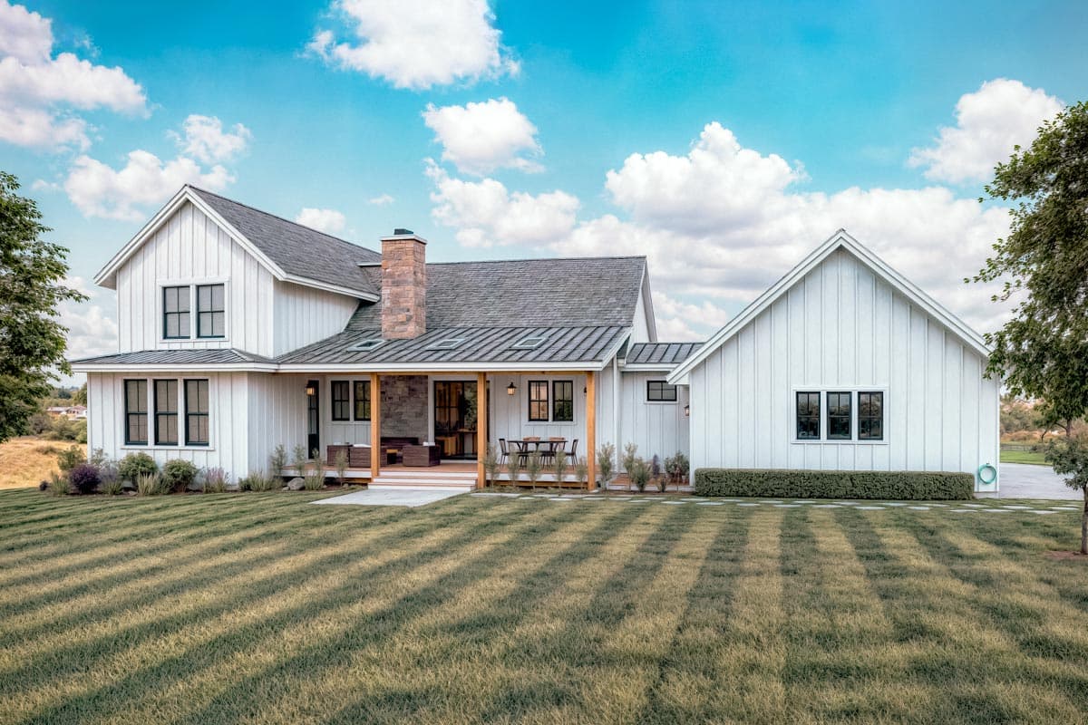 A white farmhouse with a long porch, chimney, and dark-framed windows. The house sits on a green lawn with a blue sky background.