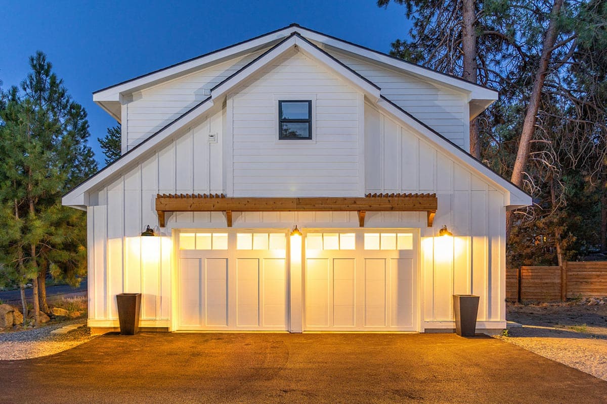 Evening shot of a two-story white house with a two-car garage. Warm lights illuminate the doors and surrounding asphalt driveway.