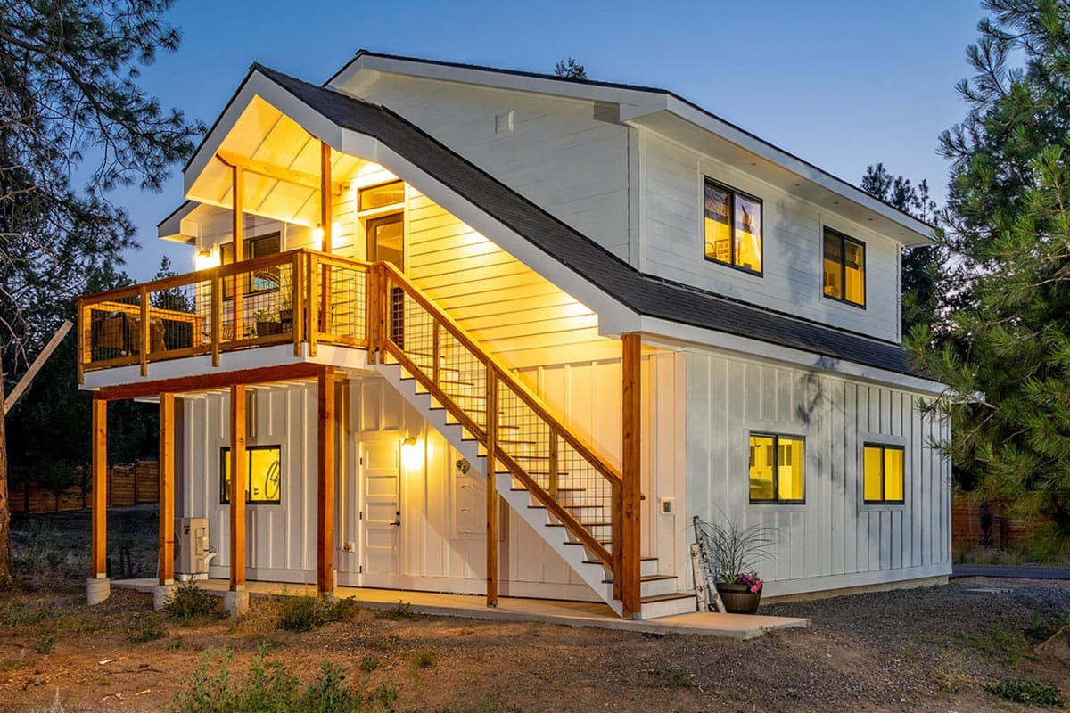 A modern two-story white house with a balcony and outdoor staircase, illuminated by warm lights against a twilight sky.