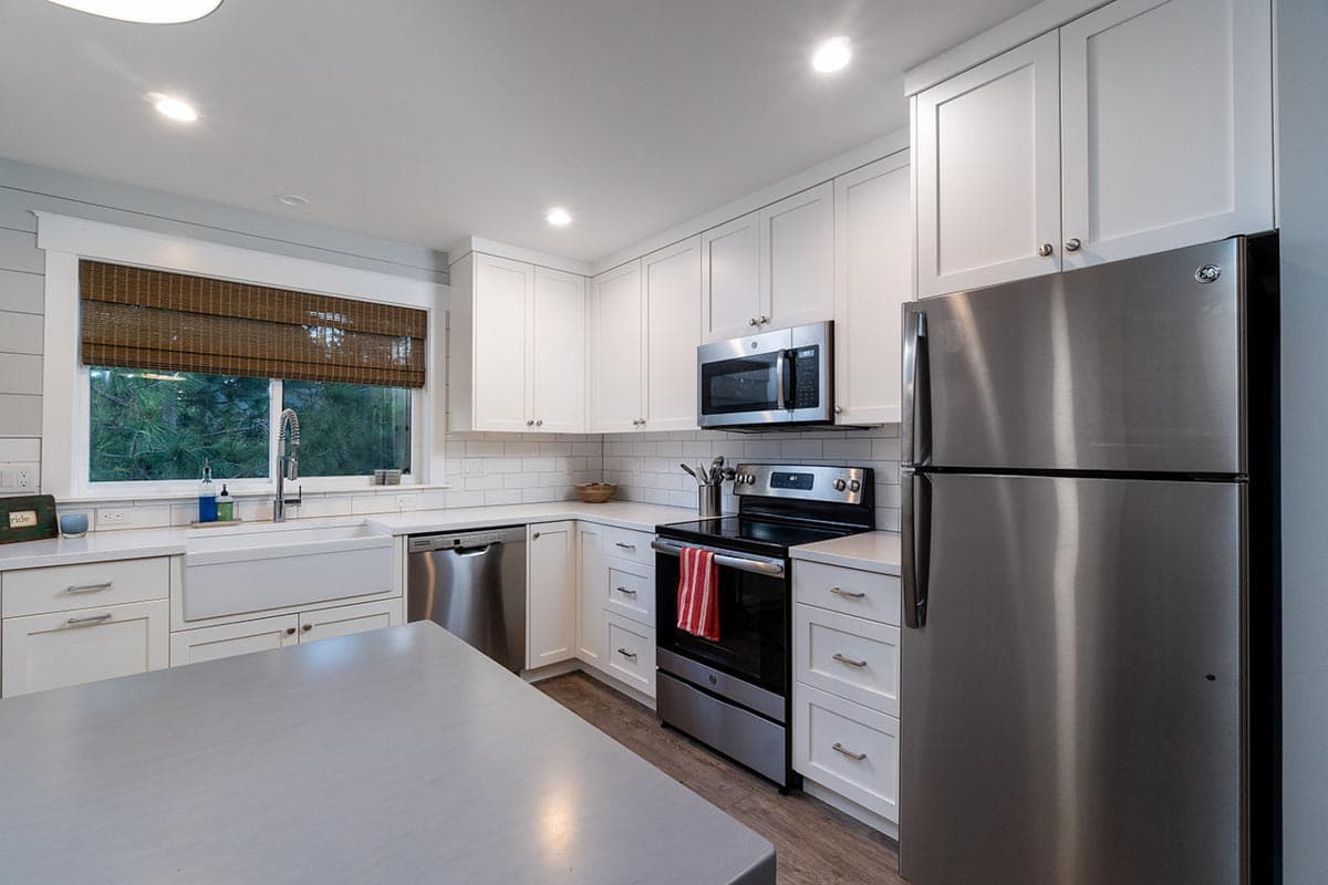 A modern kitchen with white cabinets, stainless steel appliances, and a window with a woven shade. The scene is well-lit and clean.