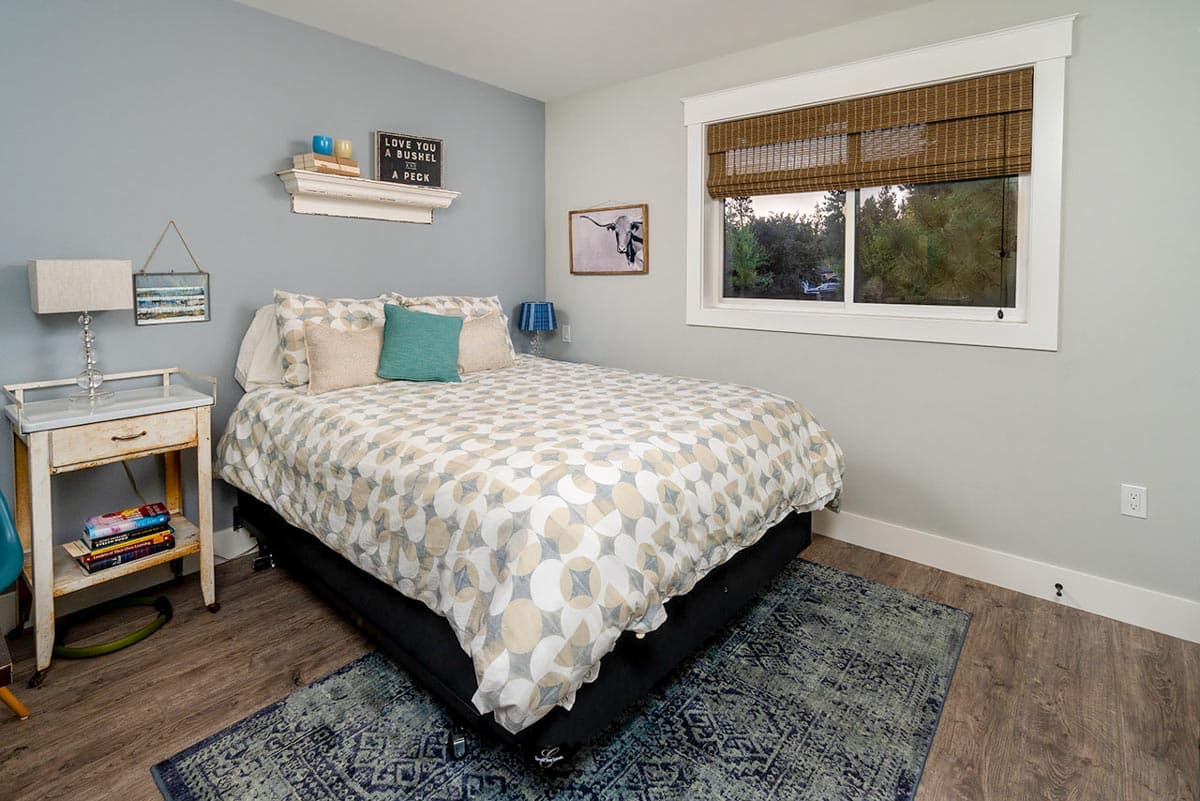 Bedroom interior featuring a double bed with patterned bedding, a nightstand, and a window with a woven shade overlooking greenery.