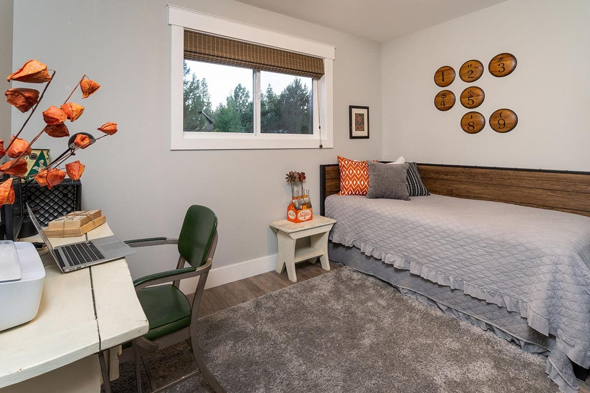 A bedroom features a bed with a gray comforter, a desk with laptop, and decor like wooden numbers on the wall. A window overlooks a forest.