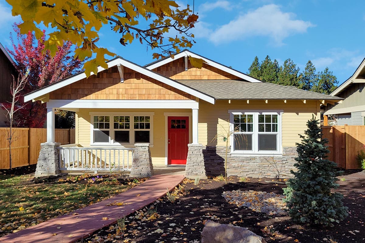 A craftsman-style house with a red front door, yellow siding, and a covered porch. Autumn leaves frame the blue sky.