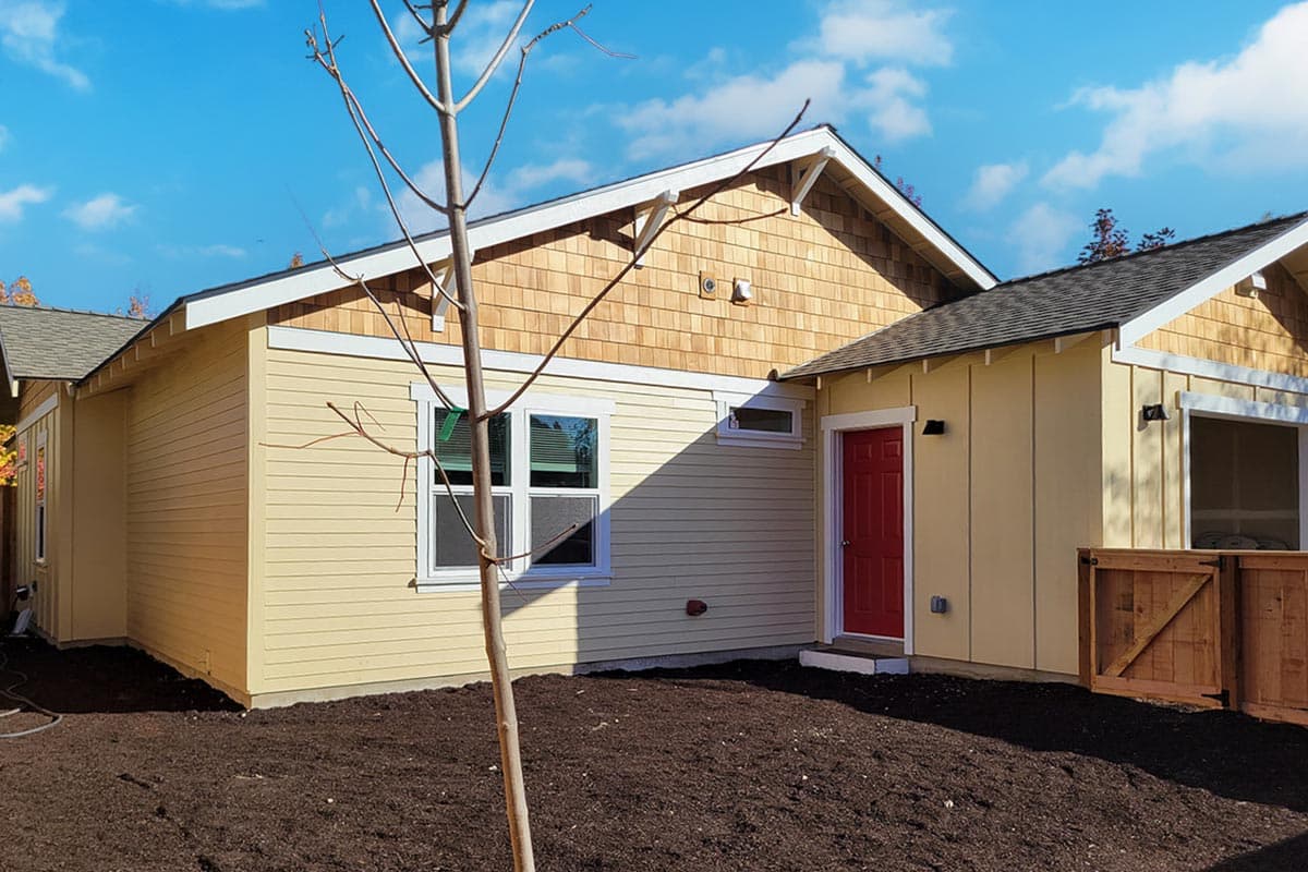 Exterior of a new construction house. The light yellow home features cedar shake siding on a gable roof and a bright red door.