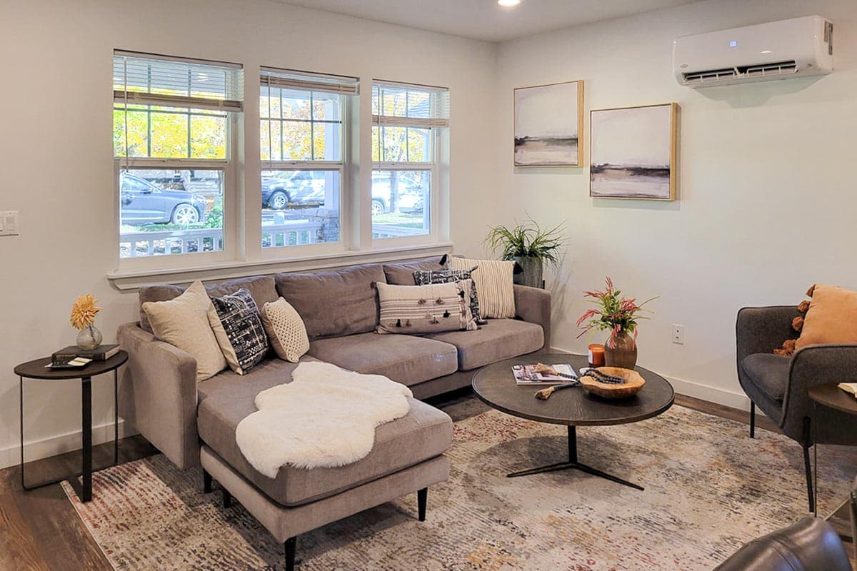 Living room with a gray sectional, round coffee table, and an area rug. Three windows reveal autumn trees and cars. Artwork adorns the walls.
