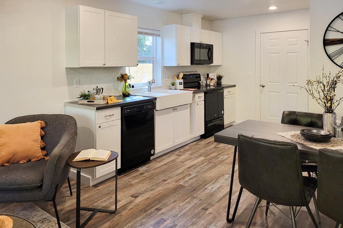 A modern kitchen with white cabinets, black appliances, and a farmhouse sink. A table and chairs sit near a wall clock.
