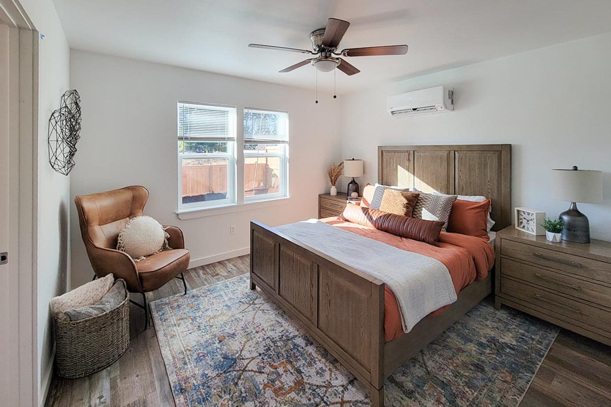 A bedroom features a wooden bed with orange bedding, a leather chair, a patterned rug, and a ceiling fan. Natural light fills the room.