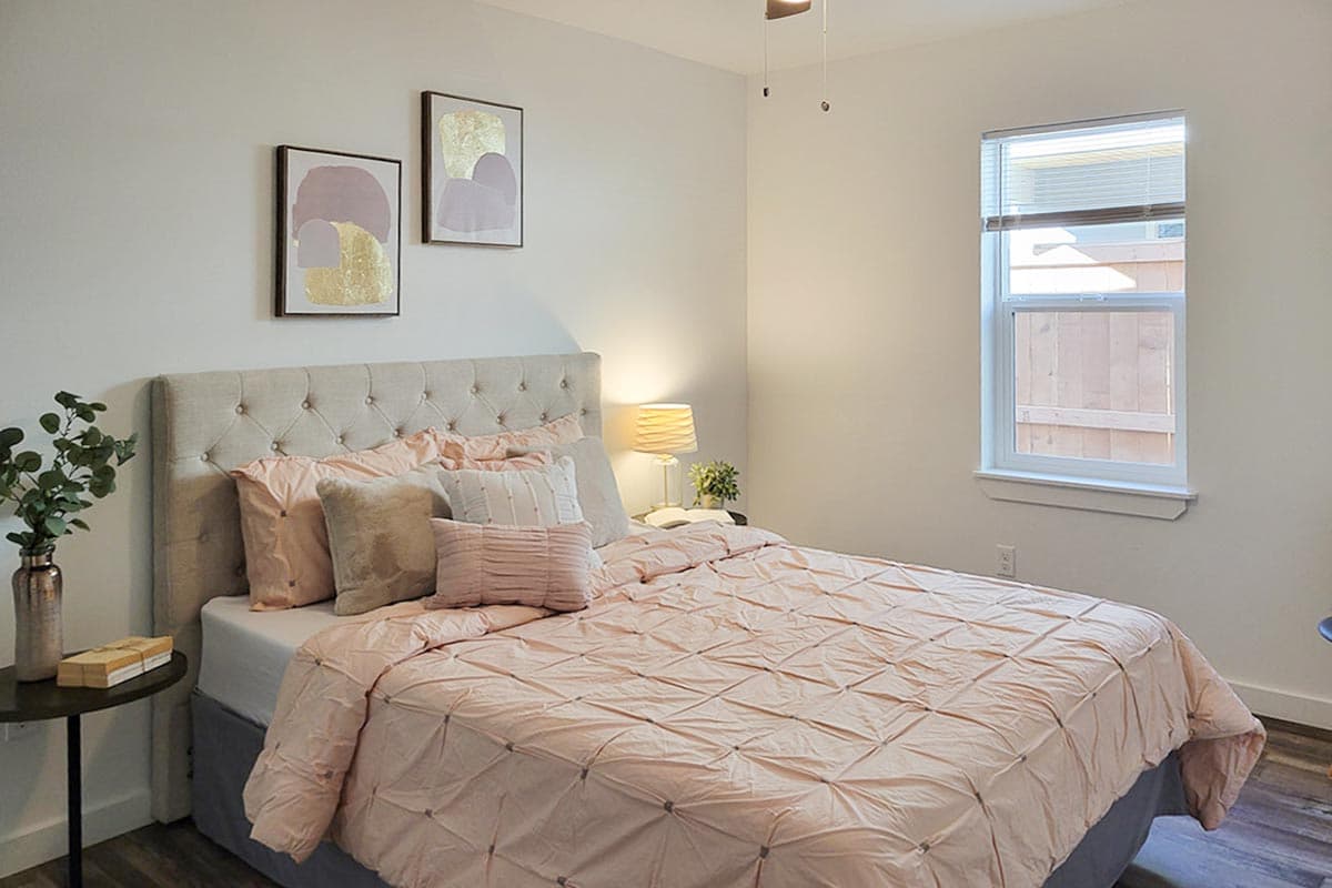 A cozy bedroom is decorated in soft pinks and creams. A tufted headboard accents the bed, which is covered with a blush comforter and pillows.