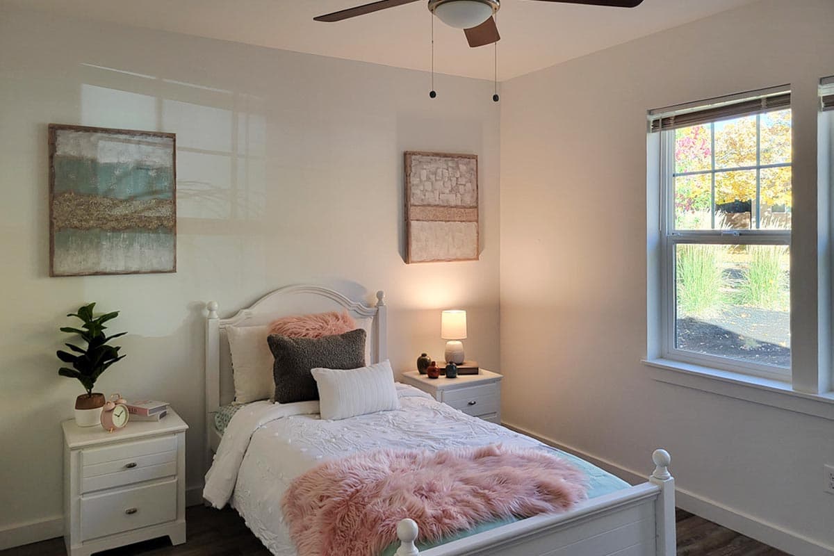 Cozy bedroom with a white bed, pink throw, and decorative pillows. A bedside table has a lamp, and artwork hangs on the wall.
