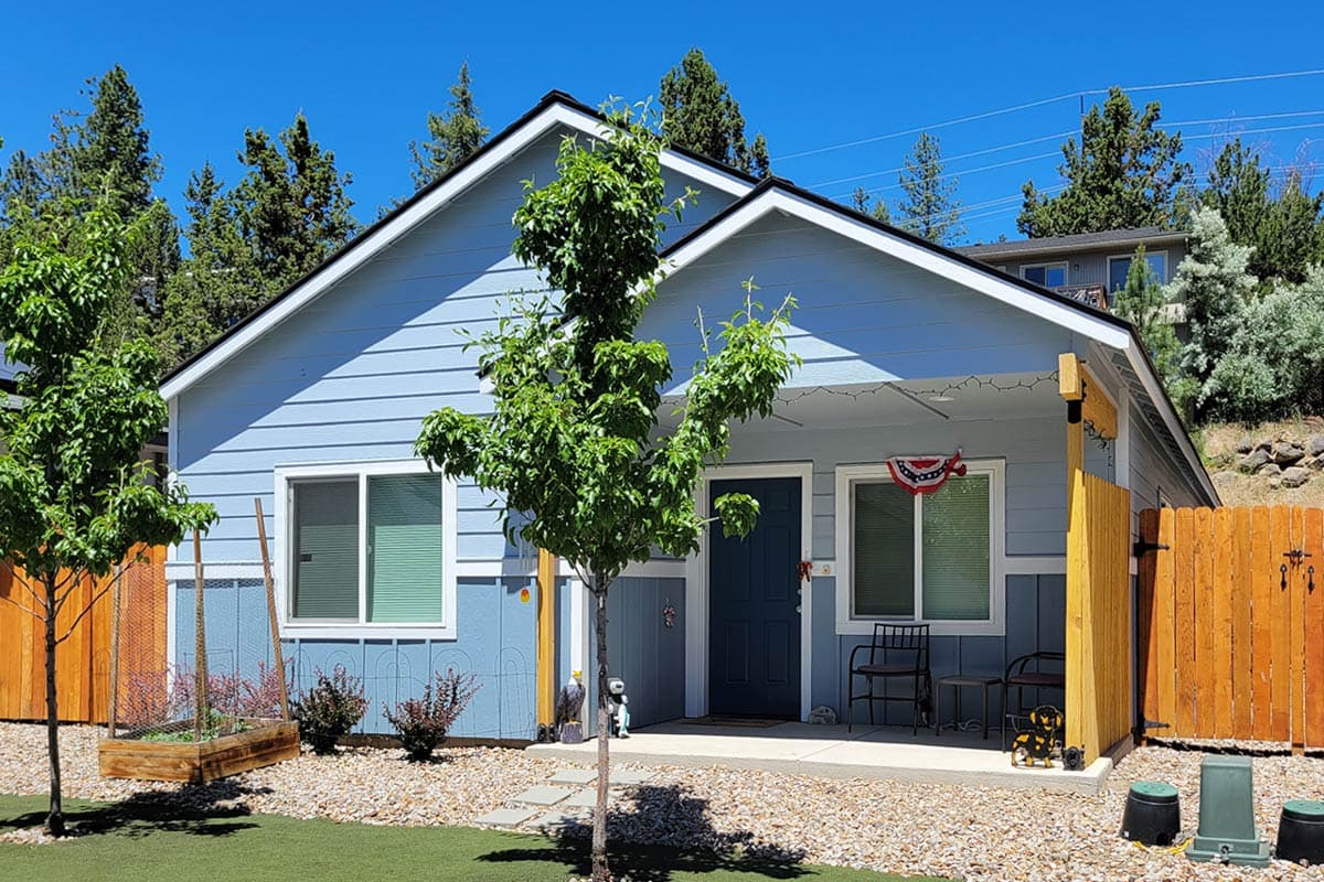 A light blue house with a small front yard is set against a backdrop of trees under a clear, blue sky. A tree shades the entrance.