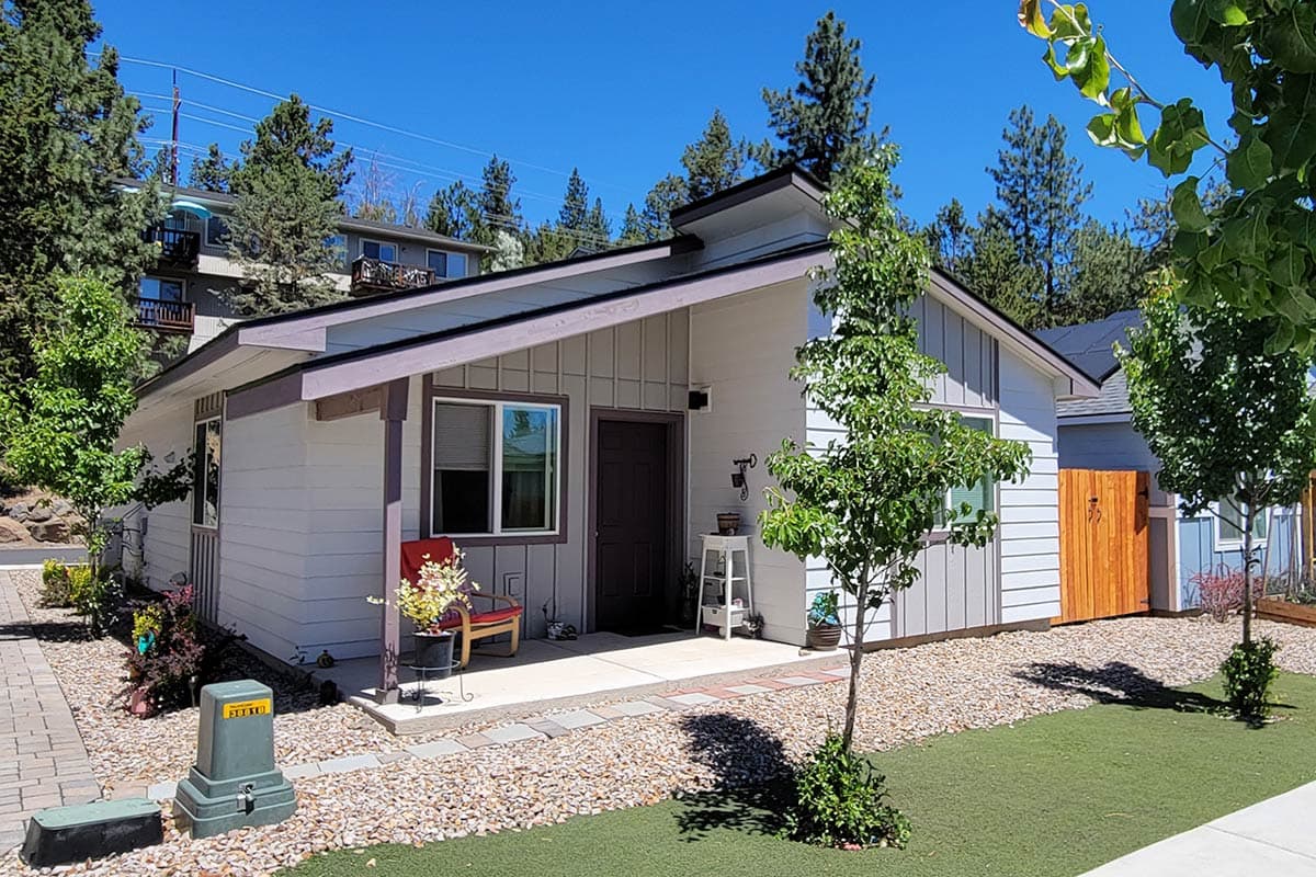 A small, light gray house with a brown door and trim sits in front of a blue sky and trees. A red chair with flowers decorates the porch.