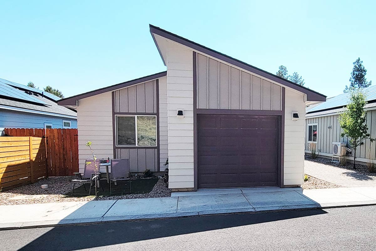 A modern one-story home with a distinctive angled roof. It has a garage door, a small yard with seating, and adjacent homes with solar panels.