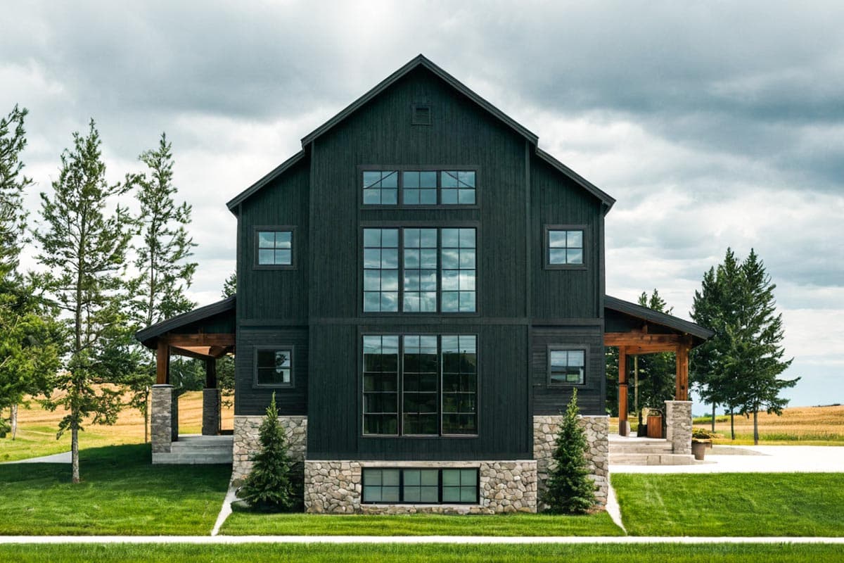 Modern farmhouse house plan exterior with dark siding, stone base, prominent gables, and a central bank of large windows.