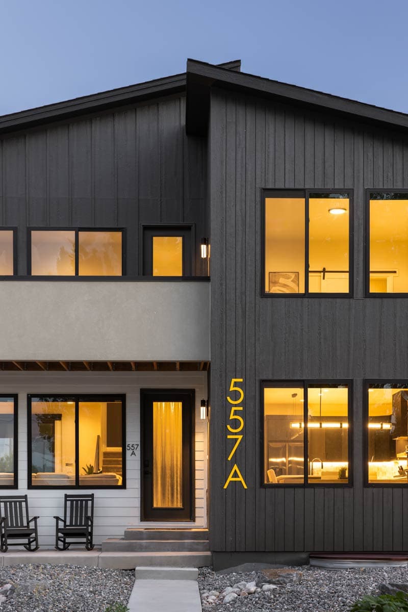 Modern house exterior with dark vertical siding, upper balcony, large windows, and prominent house numbers.