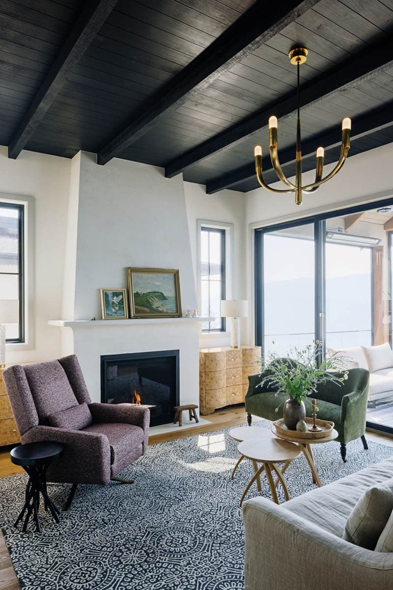 Living room with fireplace, black beamed ceiling, large windows, and patterned rug.