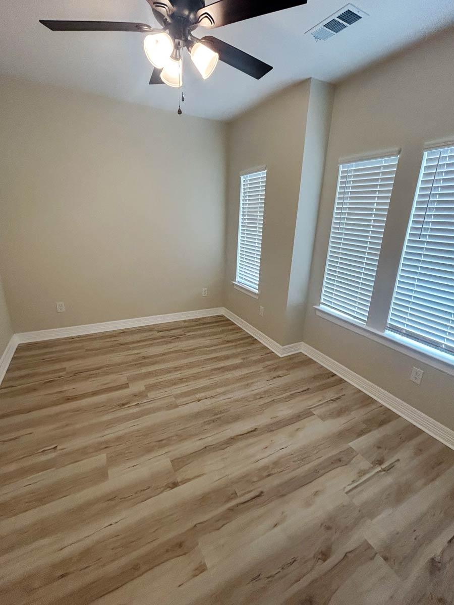 Interior view of an empty room with wood-look flooring, neutral walls, a ceiling fan, and multiple windows with blinds.