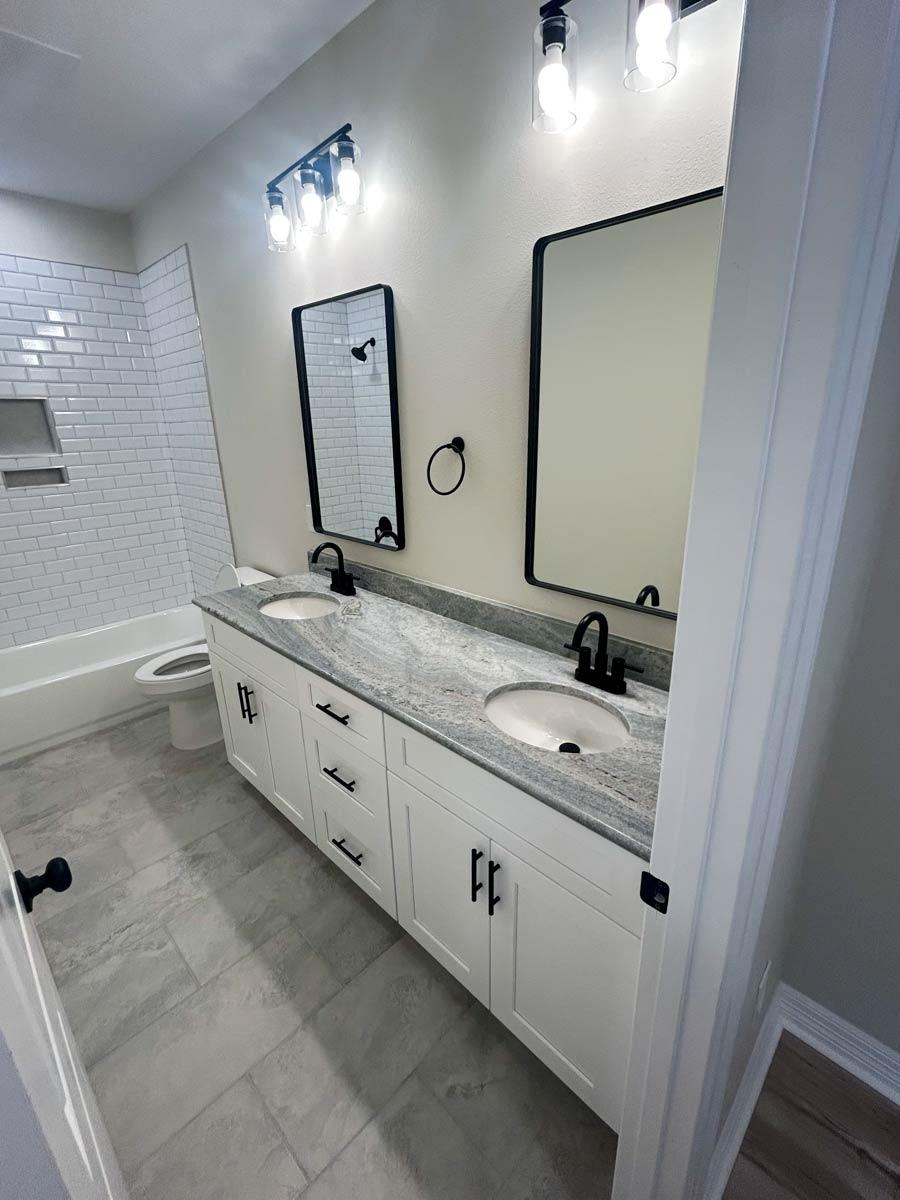 Modern bathroom with double vanity, granite countertop, subway tile shower, and gray tile flooring.